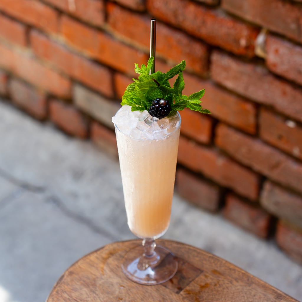 A pale peach cocktail in a tall champagne flute packed with crushed ice, garnished with fresh mint, and a blackberry
