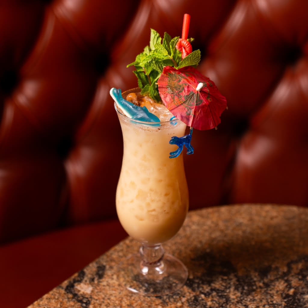 A creamy frozen colada in a hurricane glass extravagantly garnished with fresh mint, a red paper umbrella, a red straw, and a small blue plastic dinosaur