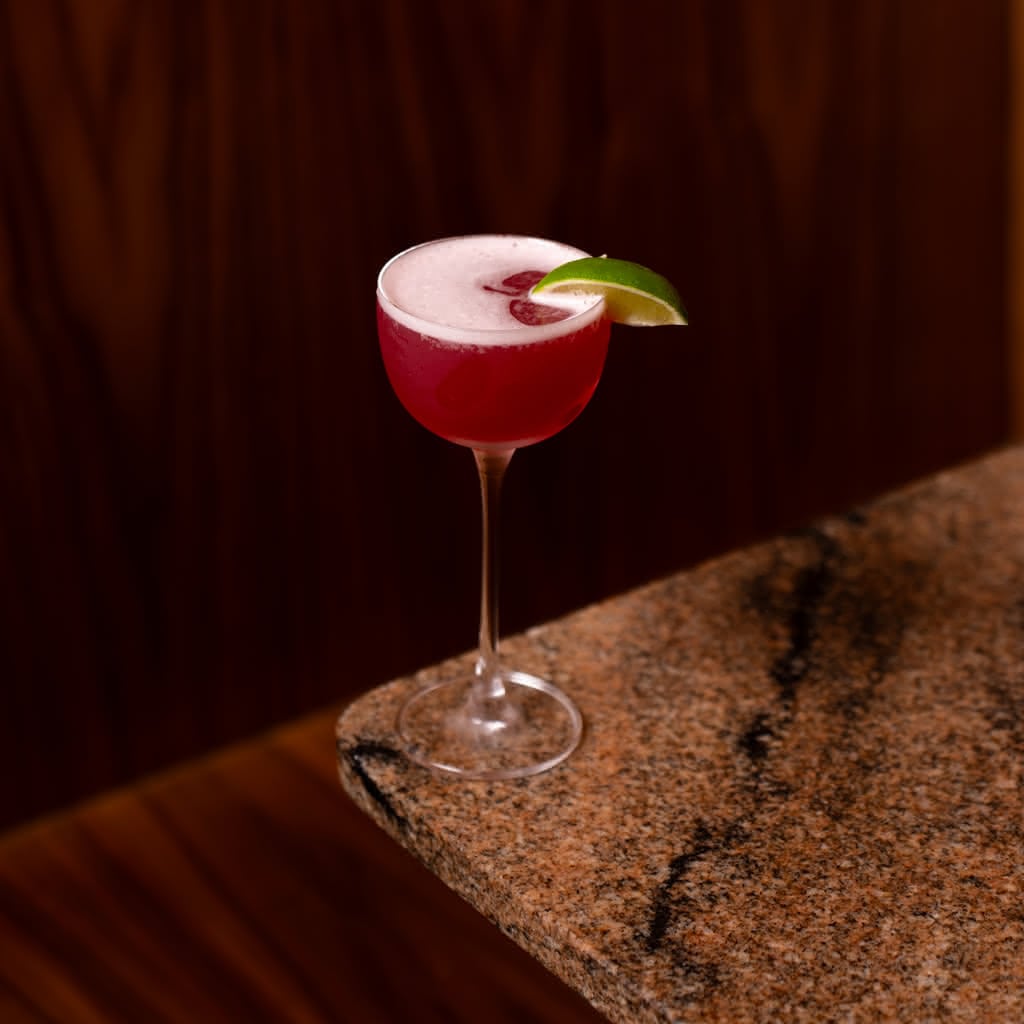 A deep crimson Cosmopolitan in a tall coupe glass with a light foam cap and lime wheel garnish