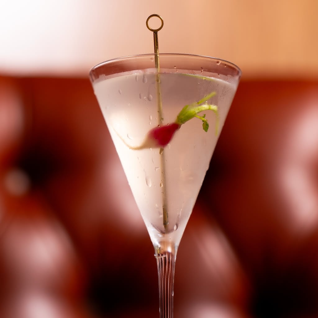 Close-up of a cone-shaped crystal martini glass holding a cloudy cocktail, garnished with a small radish and green top on a thin gold cocktail pick