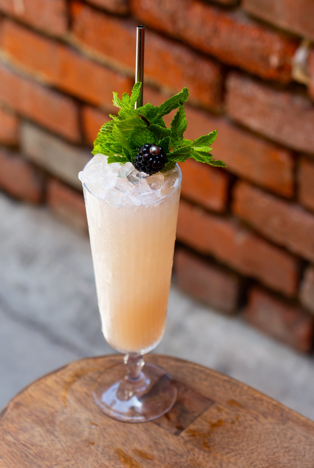 A peach-colored cocktail in a tall glass filled with crushed ice, garnished with fresh mint and a blackberry, served with a metal straw on a wooden coaster against a brick wall