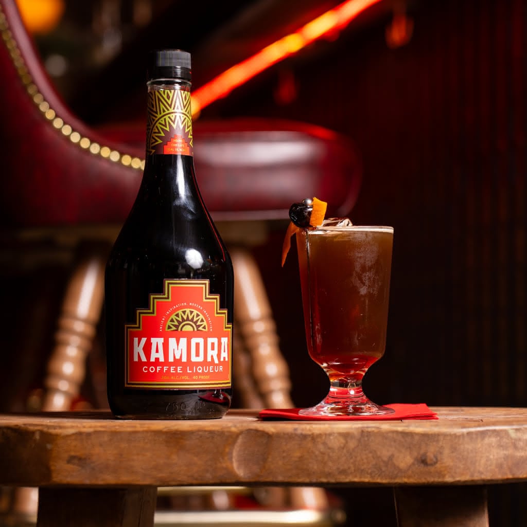 A dark cocktail in a footed glass garnished with a cherry and orange peel twist, next to a bottle of Kamora Coffee Liqueur
