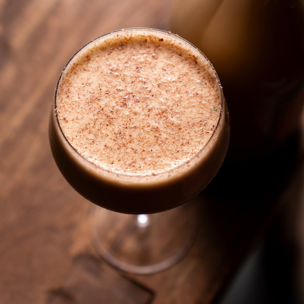 A dark cocktail in a coupe glass completely covered in thick, frothy foam dusted with grated nutmeg