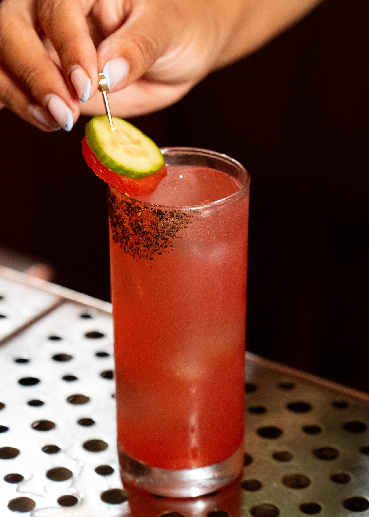 A bartender's hand placing a cucumber and tomato garnish on a pick into a tall red cocktail with a chili-salt rim, served on a perforated metal bar mat