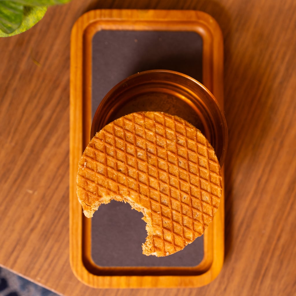 A partially eaten stroopwafel resting on top of a lowball glass on a rectangular wooden tray