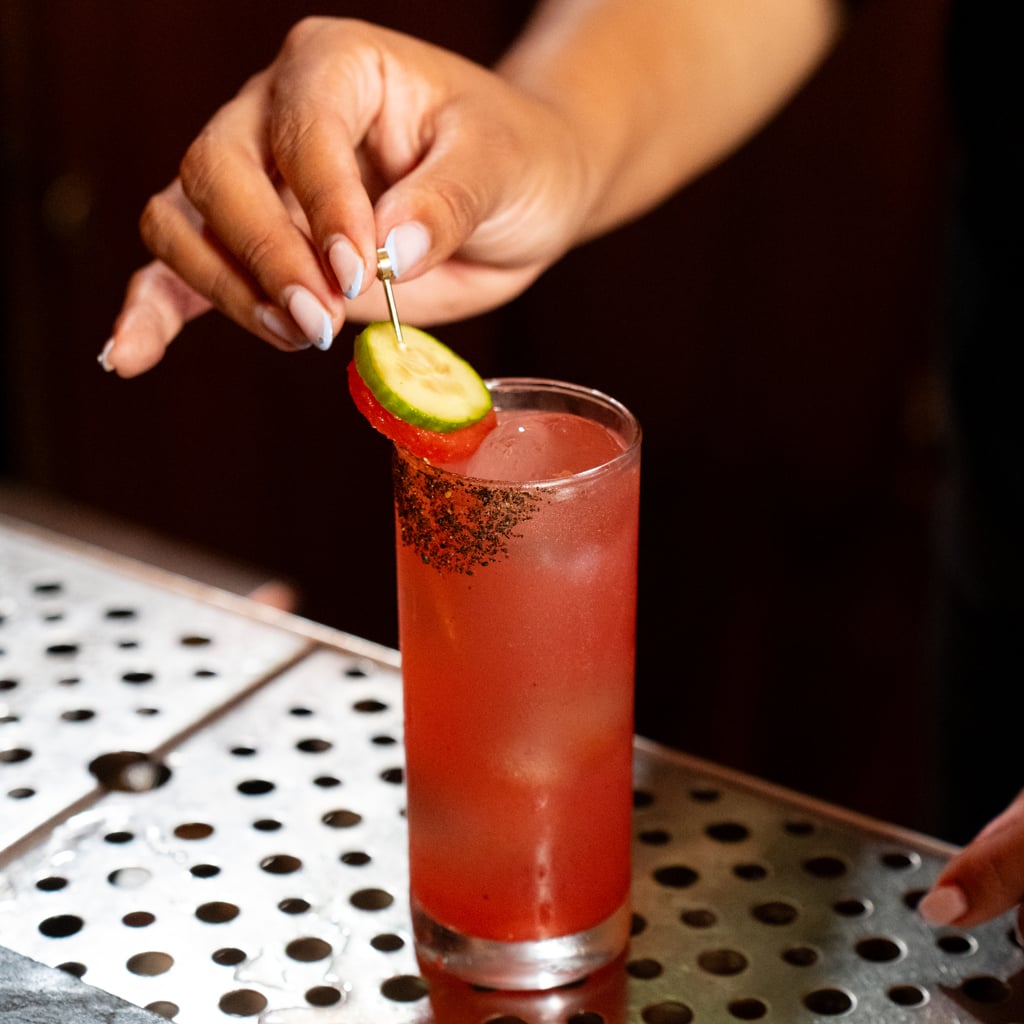 A cucumber and tomato skewer garnish a tall glass filled with a bright red cocktail with a spiced dark rim, set on a metal bar tray