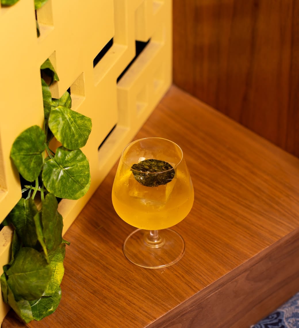 A golden Negroni in a brandy snifter with tea leaf garnish, sitting beside a slatted wall