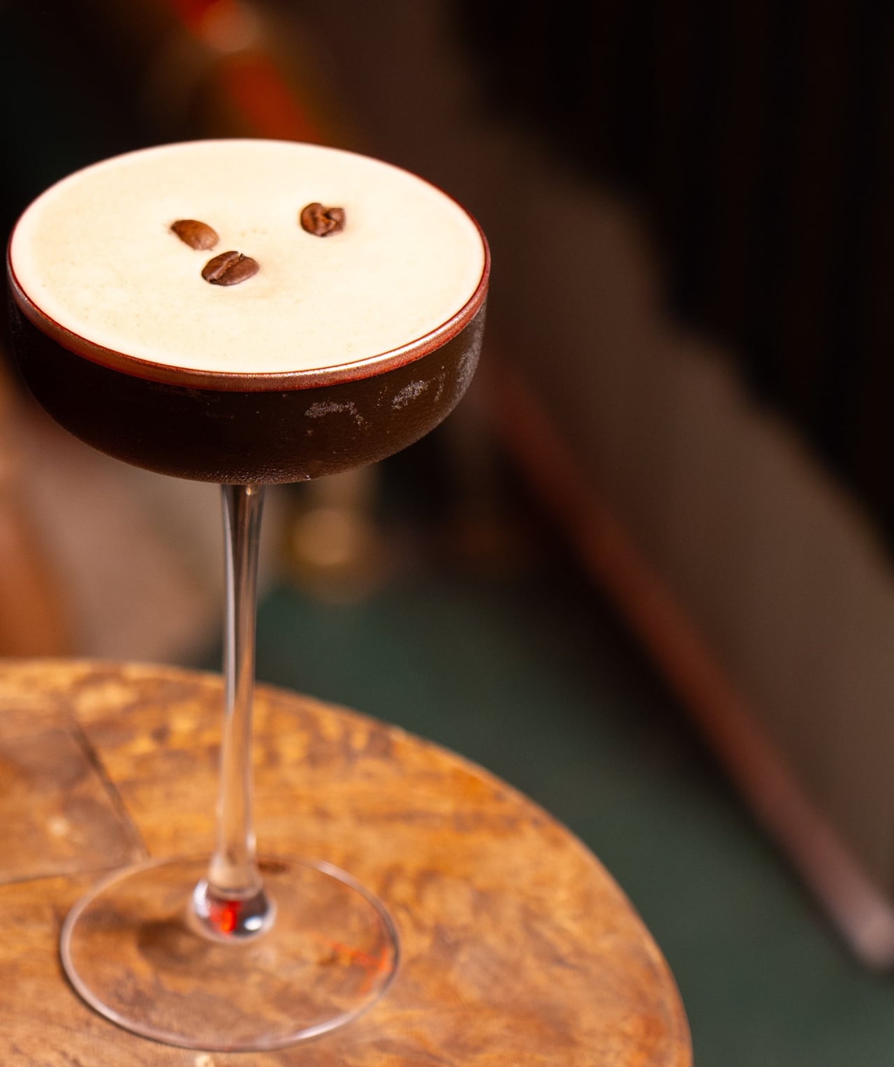 An espresso martini in a coupe glass with a thick cream foam and three coffee beans on top, resting on a wooden surface against a dark blurred background
