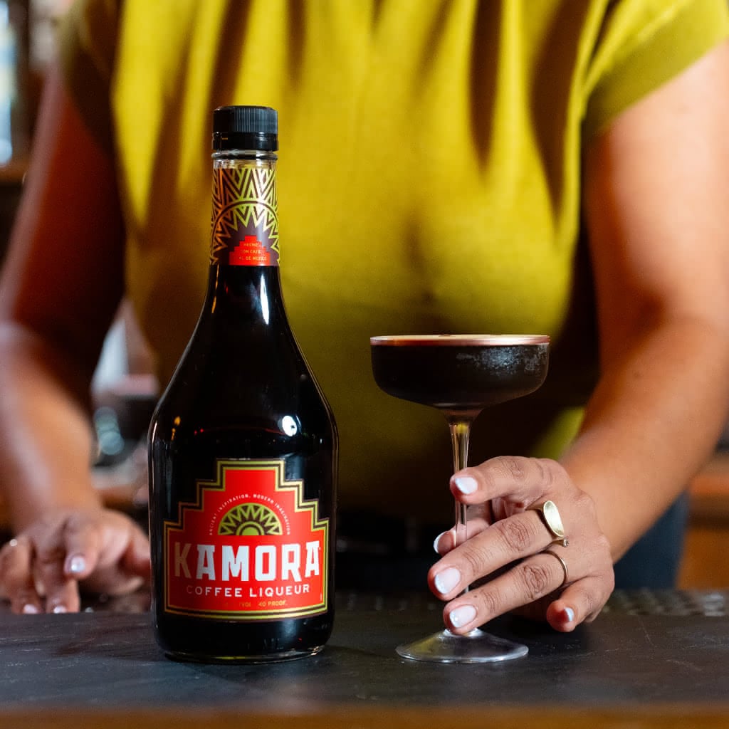 A dark espresso martini in a coupe glass next to a bottle of Kamora Coffee Liqueur against a blurred warm background