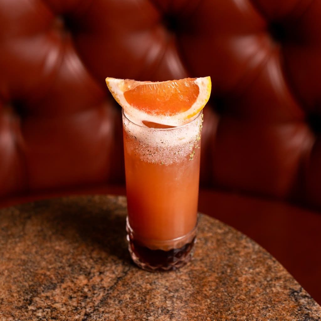 A tall ruby-red cocktail in a cut crystal highball glass, topped with foam and a spice-dusted rim, garnished with a large grapefruit wedge