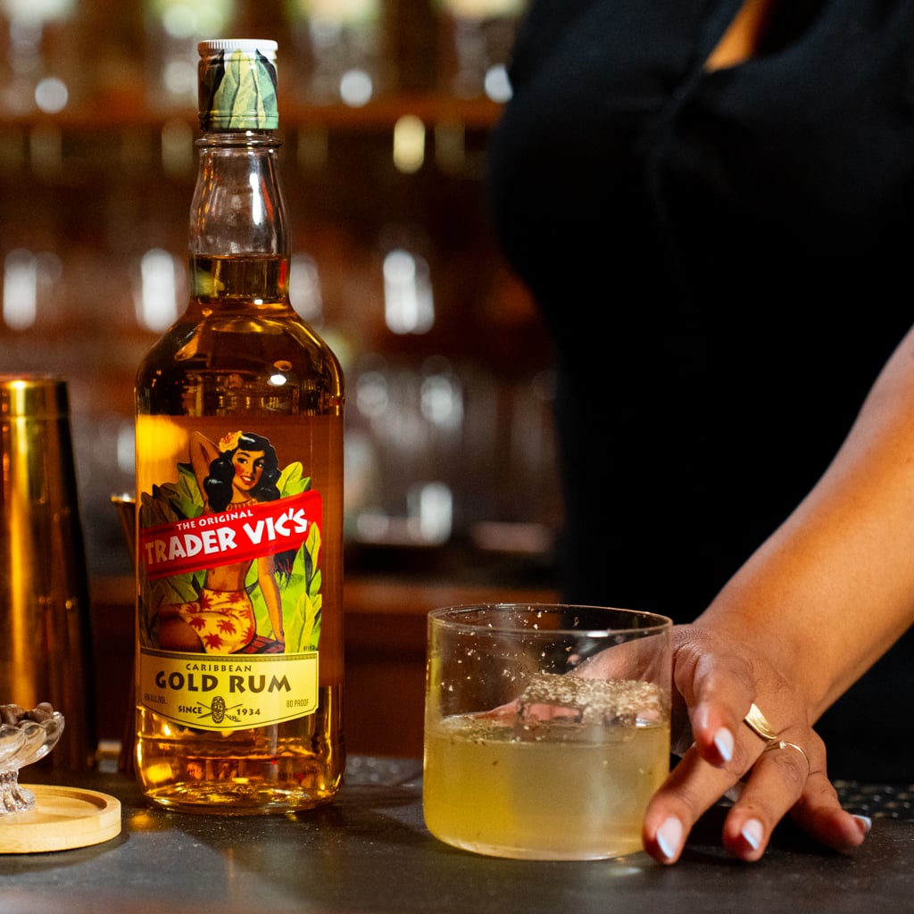 A hand reaching for a lowball glass filled with pale golden punch over ice, next to a bottle of Trader Vic's Caribbean Gold Rum