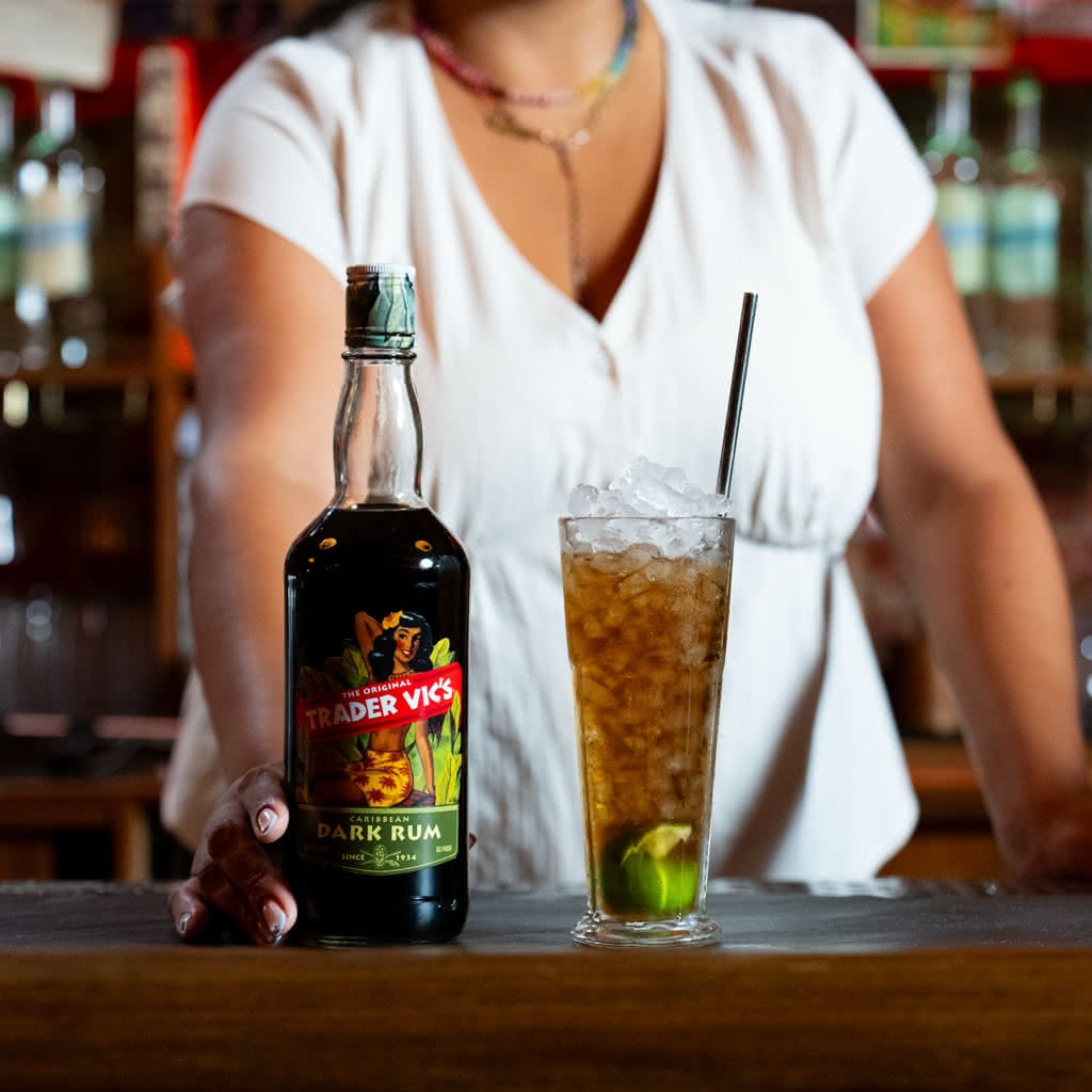 A bottle of Trader Vic's Caribbean Dark Rum beside a tall iced cola drink with a lime wedge and metal straw