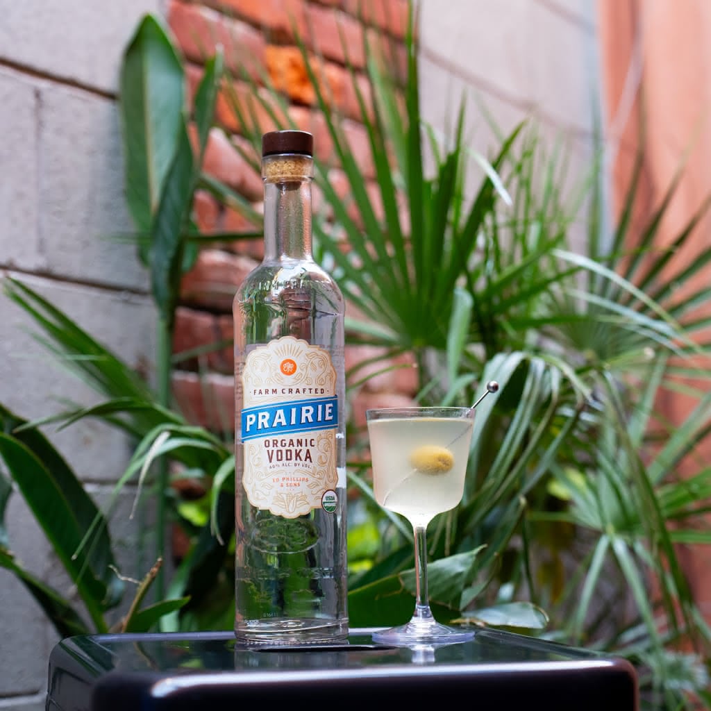A bottle of Prairie Organic Vodka next to a pale yellow-green cocktail in a coupe glass with an olive on a cocktail pin