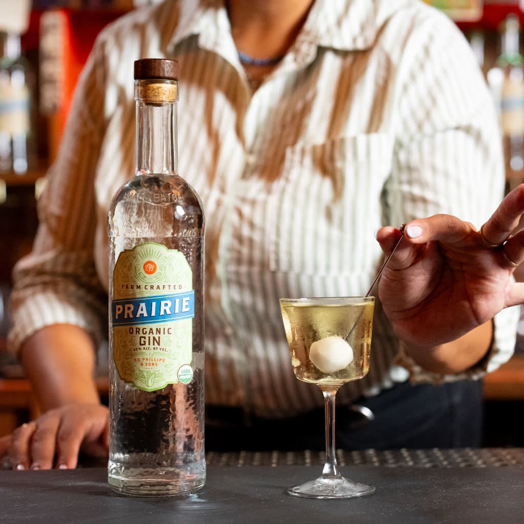 A bartender drops a white cocktail onion on a pin into a cloudy martini in a coupe glass, next to a bottle of Prairie Organic Gin