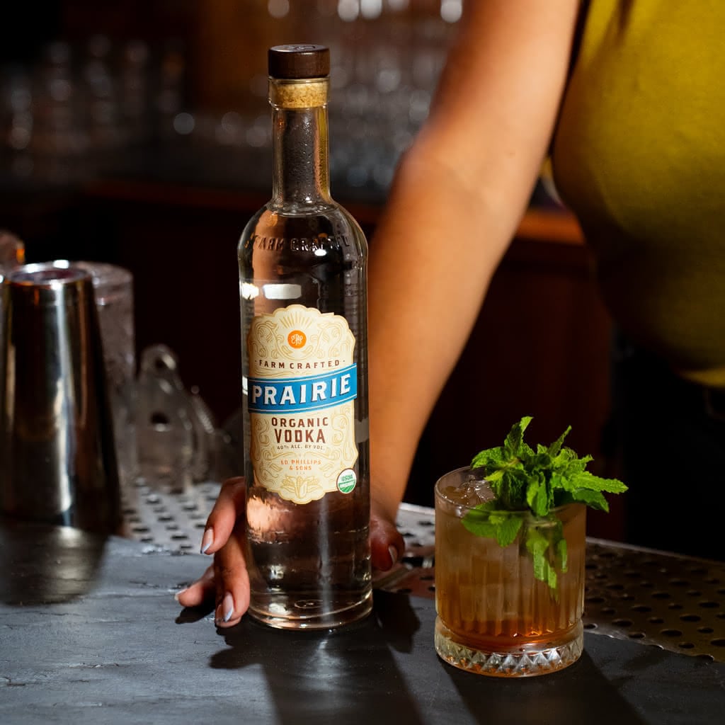 A bottle of Prairie Organic Vodka beside a lowball glass filled with an amber cocktail garnished with fresh mint, on a perforated metal bar top